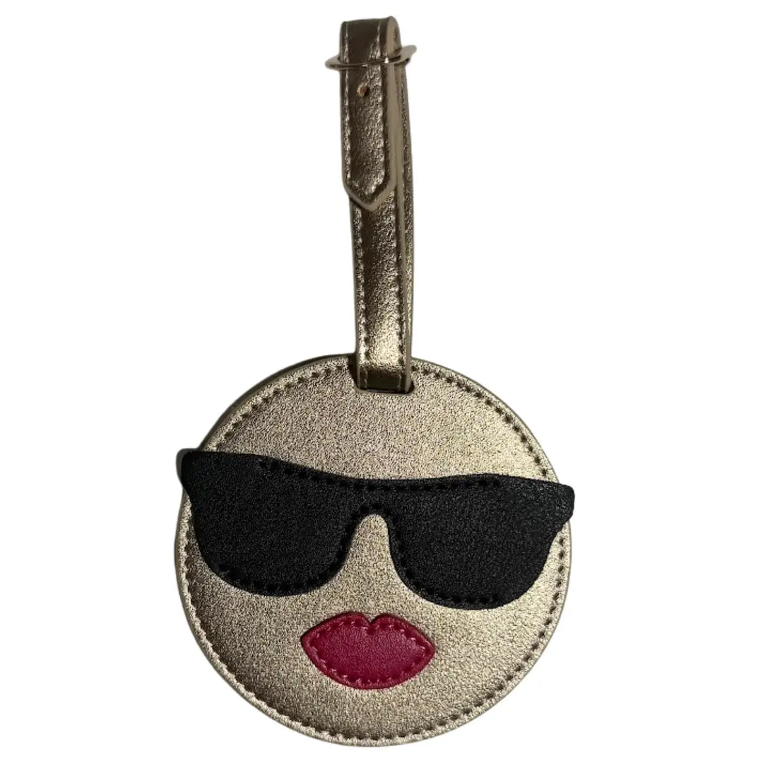 A round metallic gold luggage tag featuring an applique design of a face wearing black sunglasses and pink lips, with a gold leather strap at the top.