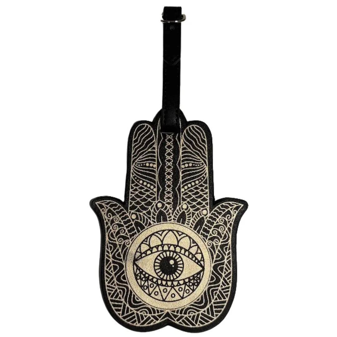 A black and cream Hamsa hand luggage tag with intricate geometric patterns and a central eye motif, featuring a black strap at the top.