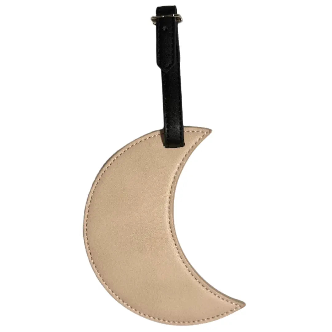 A crescent moon-shaped luggage tag in beige faux leather with a black leather strap and silver metal buckle, shown against a plain white background.