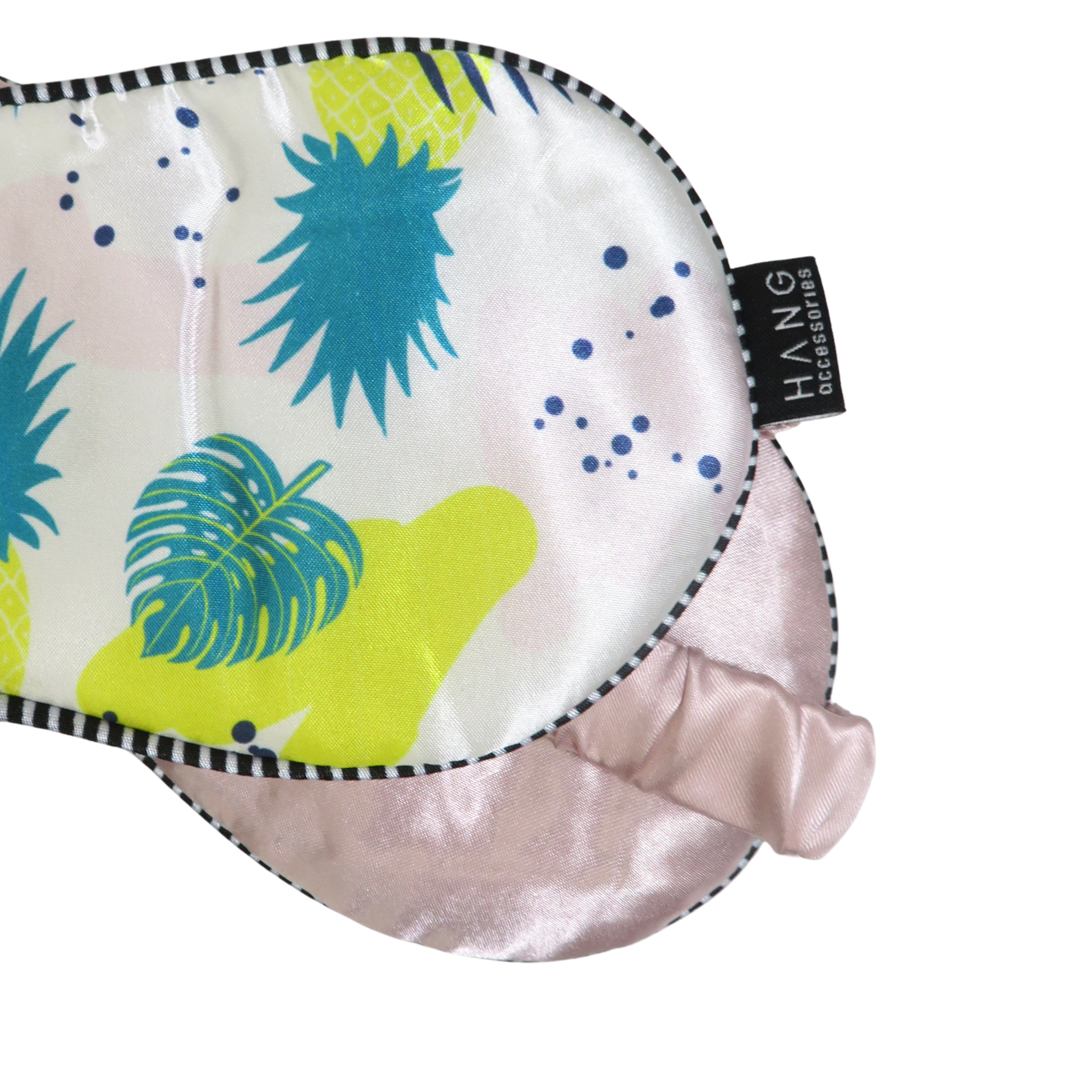 A close-up of a sleep mask with a tropical pineapple and palm leaf print on a white background, featuring a light pink satin underside and a black-and-white striped edge.