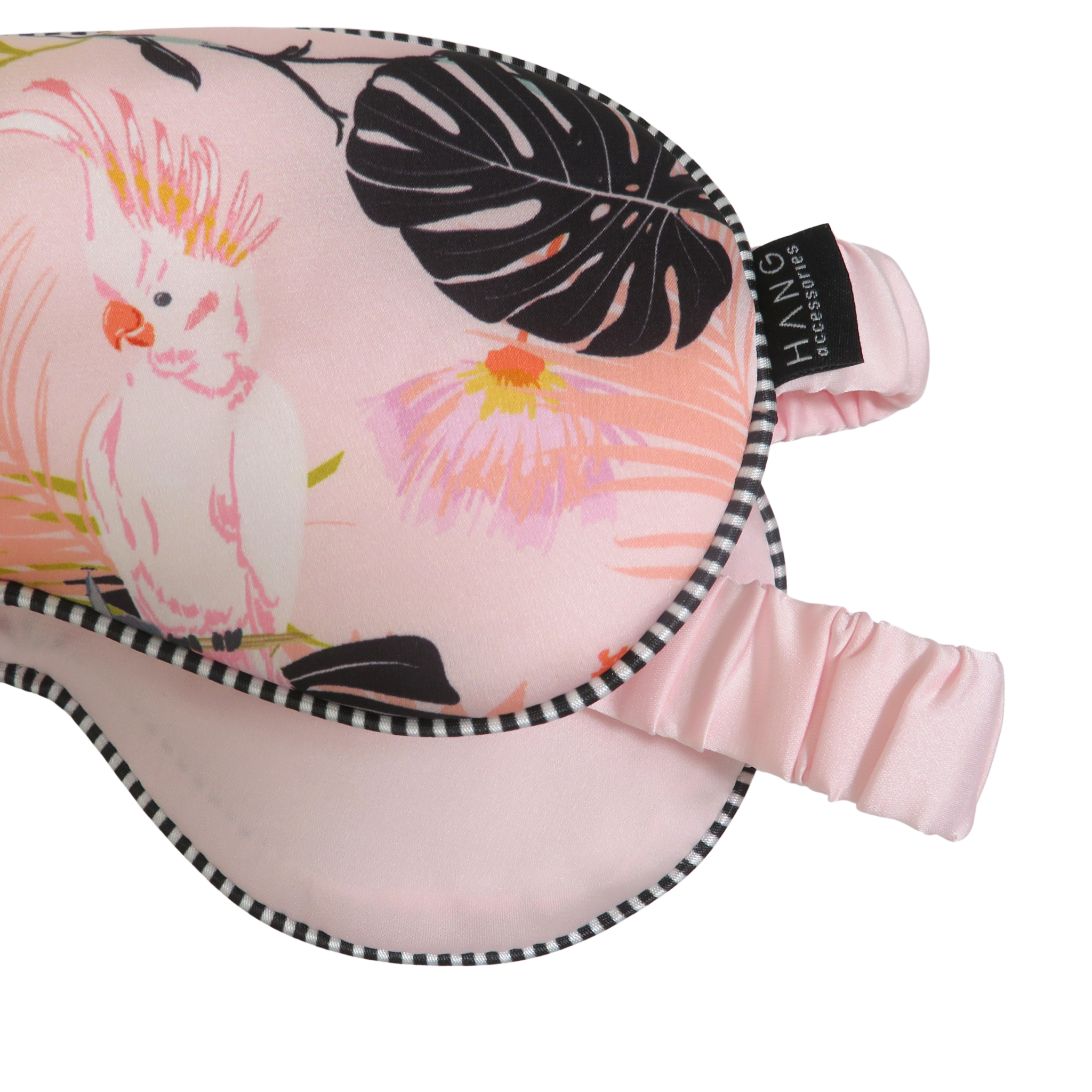 A close-up of a pink sleep mask with a tropical print featuring a cockatoo, monstera leaves, and pink flowers. The mask has a black and white checkered trim.