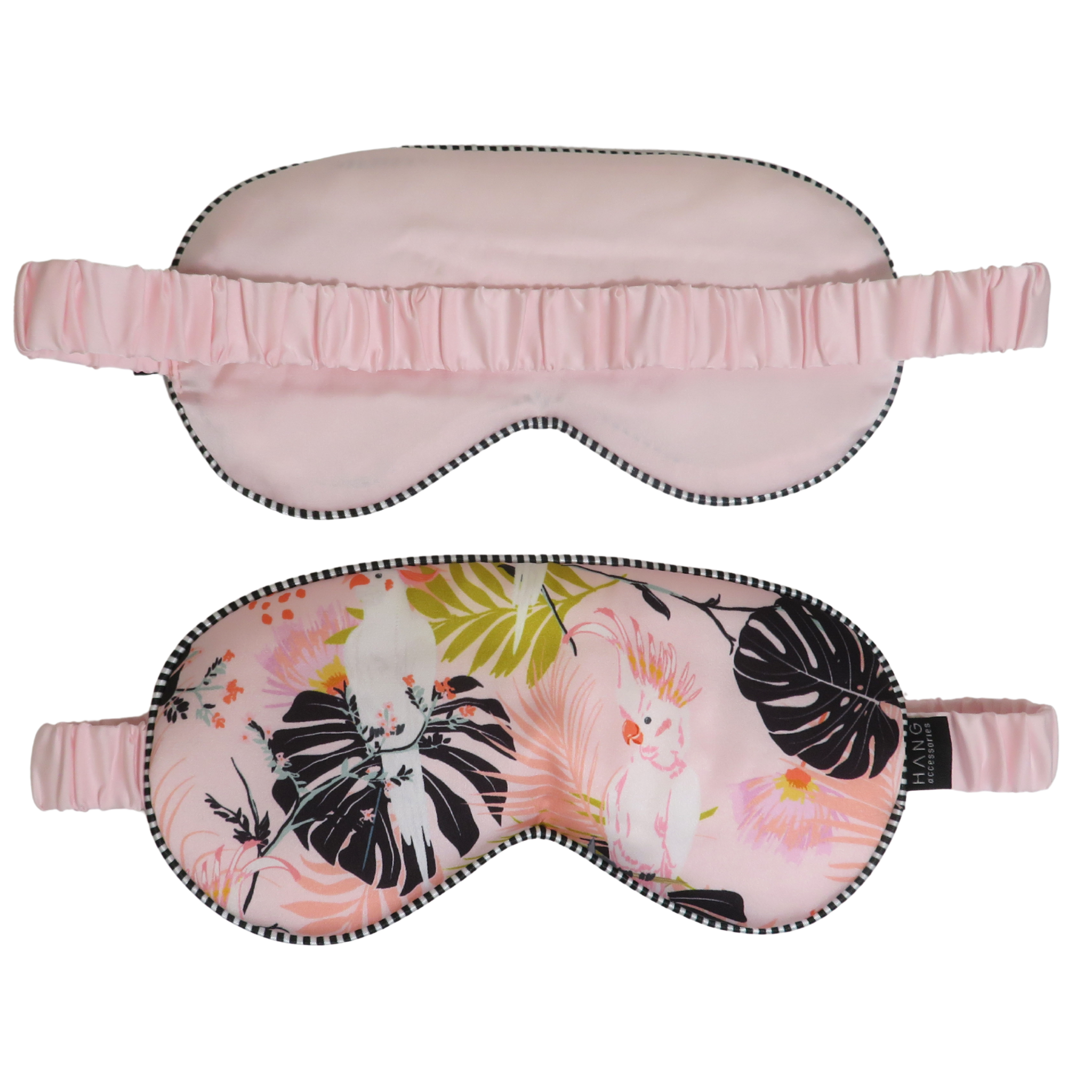 Two sleep masks shown on a plain white background: one solid light pink mask with a ruffled strap and black stitched edge, and one matching pink mask with a tropical floral print featuring black monstera leaves, pink flowers, and a white bird, also with a ruffled pink strap and black stitched edge.