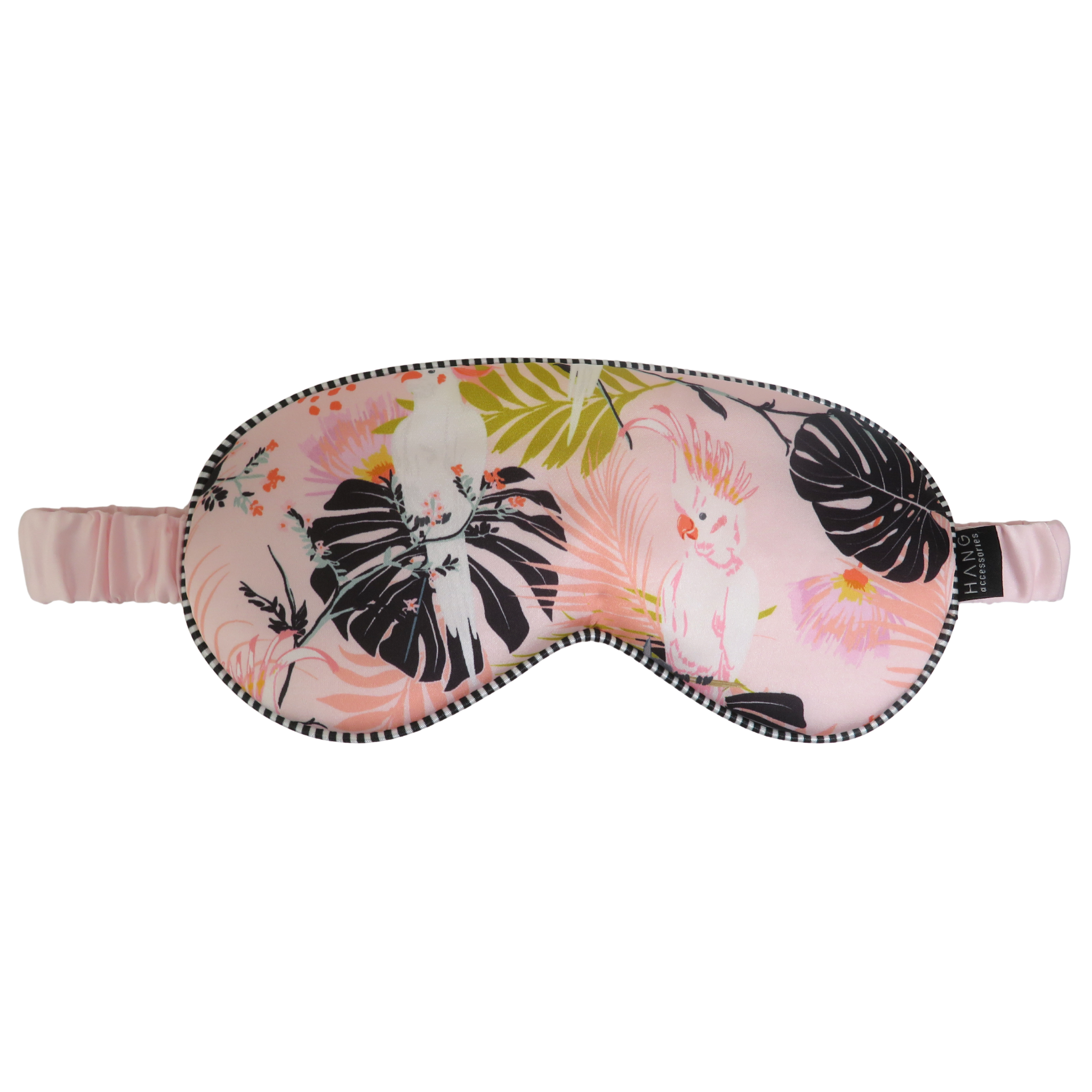 A pink satin sleep mask with a tropical print featuring black monstera leaves, cockatoos, and green palm leaves, complete with an elastic strap.