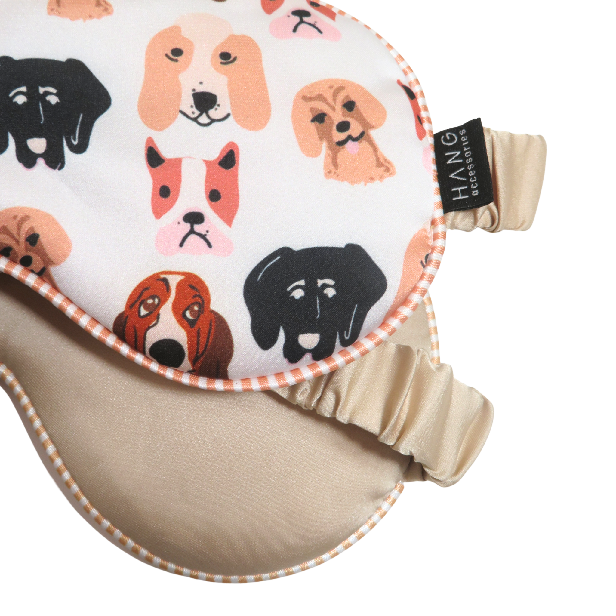 Close-up of a dog-themed eye mask with a white printed fabric featuring various dog faces in black, brown, and orange, finished with a beige satin strap.