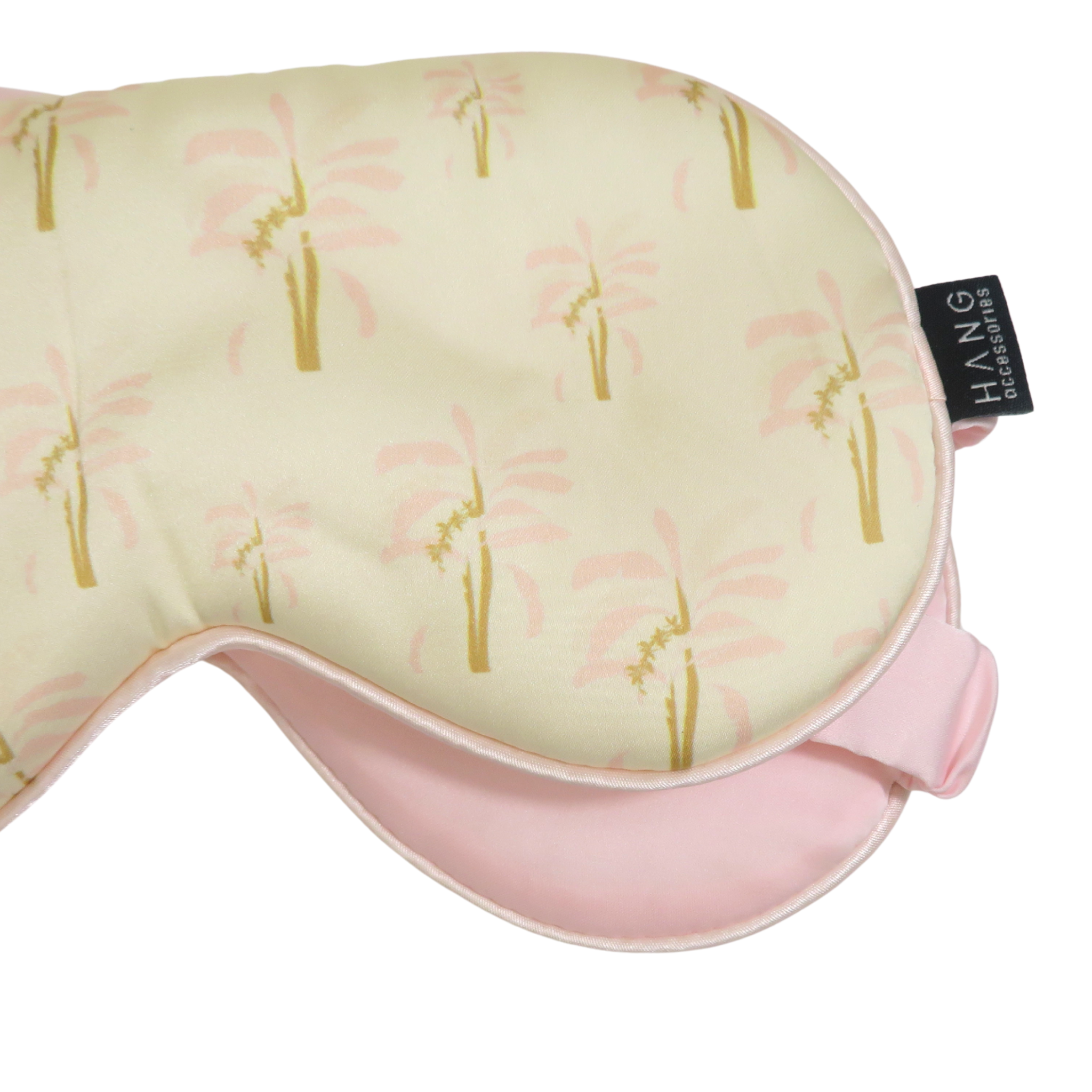 A close-up of a sleep mask with a pale yellow and pink palm tree pattern, featuring a pink underside.