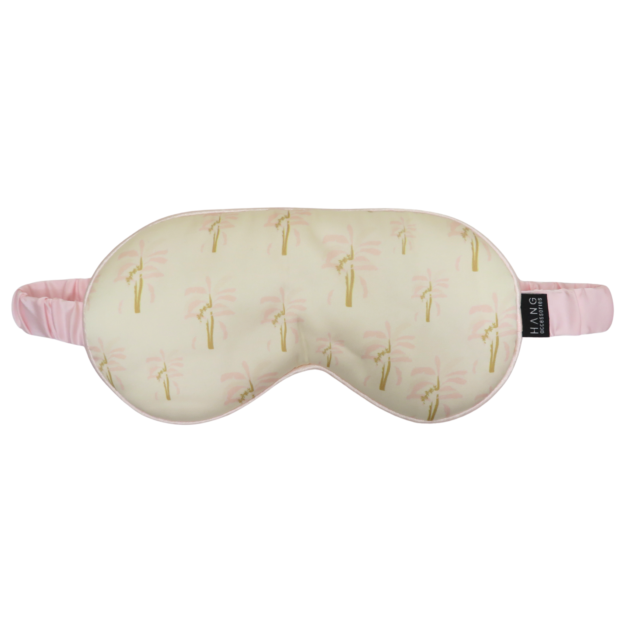 A sleep mask with a cream-colored background, a pink and gold palm tree pattern, and a pink elastic strap. 